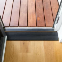 Ein schwarzer Schwellenkeil schafft einen barrierefreien Übergang zwischen einer Holzterrasse im Freien und einem hellen Holzfußboden im Inneren.