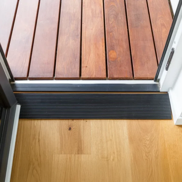 Ein schwarzer Schwellenkeil schafft einen barrierefreien Übergang zwischen einer Holzterrasse im Freien und einem hellen Holzfußboden im Inneren.
