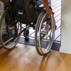 Eine Person im Rollstuhl fährt über einen Schwellenkeil mit niedriger Kante von einem Holzboden im Innenbereich auf eine Terrasse im Außenbereich.