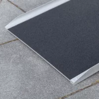 Nahaufnahme der unteren Auflagefläche einer Rollstuhlrampe mit strukturierter schwarzer Oberfläche auf einem grauen Betonpflaster.