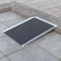 Eine tragbare Rollstuhlrampe wird über einen Bordstein auf einem gepflasterten Gehweg gelegt.