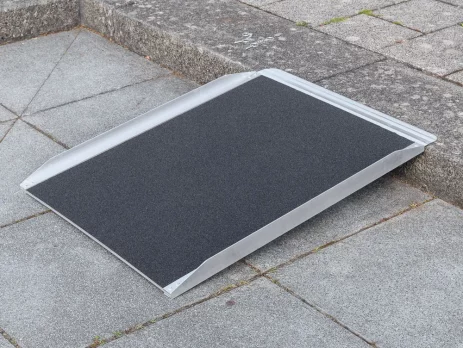 Eine tragbare Rollstuhlrampe wird über einen Bordstein auf einem gepflasterten Gehweg gelegt.