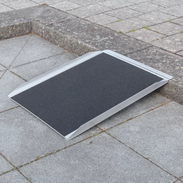 Eine tragbare Rollstuhlrampe wird über einen Bordstein auf einem gepflasterten Gehweg gelegt.