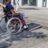 Eine Person im Rollstuhl nähert sich einer Bordsteinkante über eine tragbare Rampe auf einem gepflasterten Gehweg.