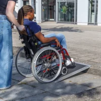 Eine Person im Rollstuhl wird an einem sonnigen Tag im Freien von einer anderen Person eine Bordsteinrampe hinaufgefahren.