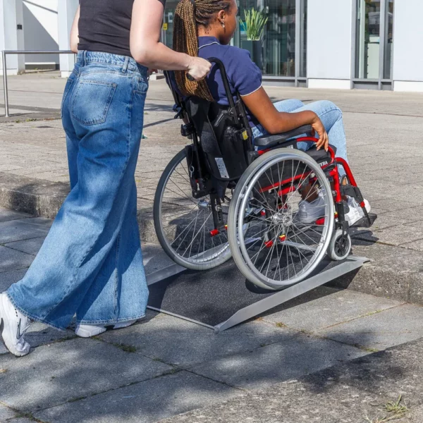 Eine Person schiebt eine andere Person im Rollstuhl über eine tragbare Rampe auf einen Bordstein vor einem Gebäude.