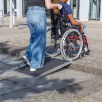 Eine Person schiebt eine andere Person im Rollstuhl auf eine tragbare Rampe über einen Bordstein auf einem städtischen Bürgersteig.