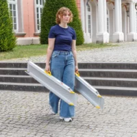 Eine Person in Jeans und marineblauem Shirt trägt zwei Rollstuhlrampen-Schienen im Freien in die Nähe von Steinstufen und einem Gebäude mit Säulen.