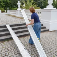 Eine Person in Jeans stellt eine tragbare Aluminiumrampe über Steinstufen im Freien in der Nähe eines weißen Geländers auf.