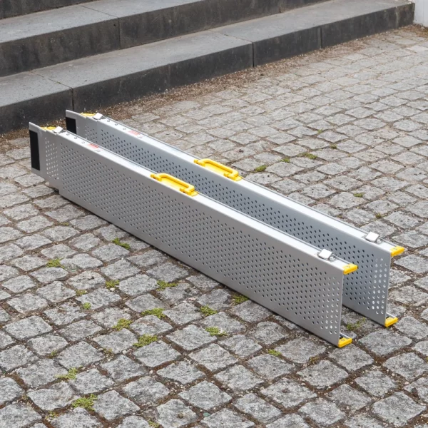 Zwei tragbare Rollstuhlrampen-Schienen mit gelben Griffen sind nebeneinander auf einem Kopfsteinpflaster in der Nähe einiger Stufen aufgestellt.