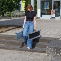 Eine Person in schwarzem Shirt und blauer Jeans trägt zwei Elemente einer Rollstuhlrampe