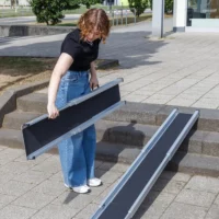 Eine Person in Jeans und schwarzem Shirt stellt eine tragbare Rampe über einer Außentreppe auf einem gepflasterten Gehweg auf.