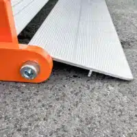 Nahaufnahme der unteren Auflage einer orangefarbenen Rollrampe mit einer Innensechskantschraube, der auf einer rauen Betonoberfläche aufliegt.