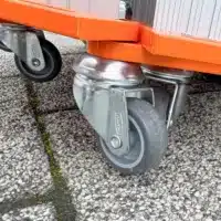 Nahaufnahme von drehbaren Metallrollen, die an eine orangefarbenen Rollrampe angebracht sind, die auf einer strukturierten Oberfläche im Freien liegt.