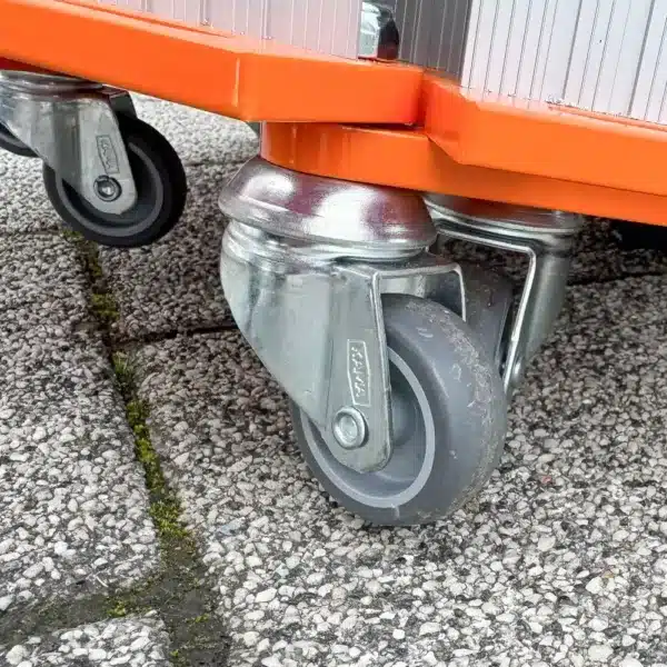 Nahaufnahme von drehbaren Metallrollen, die an eine orangefarbenen Rollrampe angebracht sind, die auf einer strukturierten Oberfläche im Freien liegt.