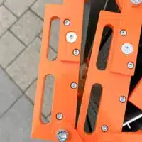Nahaufnahme der seitlichen Aluminiumelemente einer orangefarbenen Rollrampe mit Innensechskantschrauben, die über grauen Pflastersteinen im Hintergrund liegt.