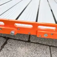 Detailaufnahme der orangefarbenen Seitenelemente einer Rollrampe mit aluminiumfarbener Fahrfläche, die auf einer Steinfliesenfläche liegen.