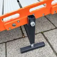 Ein schwarzer Standfuss aus Metall, befestigt am Seitenteil einer orangefarbenen Rollrampe, welcher auf einer strukturierten Gehwegoberfläche steht.