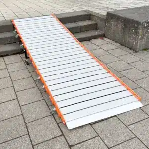 Eine trag- und rollbare Rollstuhlrampe aus Metall mit orangefarbenen Seitenteilen wird über Betonstufen auf einen gepflasterten Gehweg gelegt.