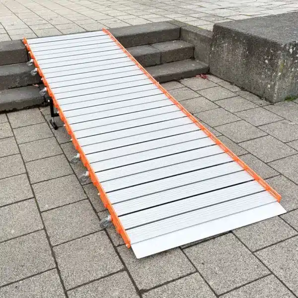 Eine trag- und rollbare Rollstuhlrampe aus Metall mit orangefarbenen Seitenteilen wird über Betonstufen auf einen gepflasterten Gehweg gelegt.