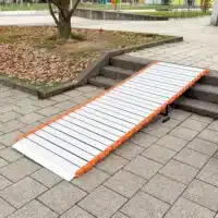 Eine tragbare Metallrampe mit orangefarbenen Seitenteilen ist über Stufen auf einem gepflasterten Gehweg in der Nähe eines Baumes angebracht.