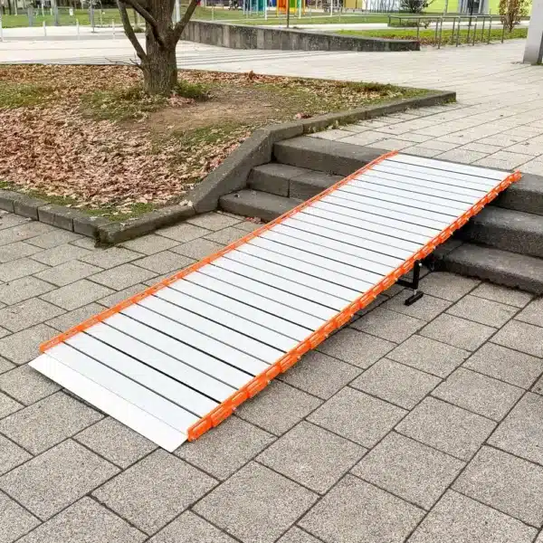 Eine tragbare Metallrampe mit orangefarbenen Seitenteilen ist über Stufen auf einem gepflasterten Gehweg in der Nähe eines Baumes angebracht.