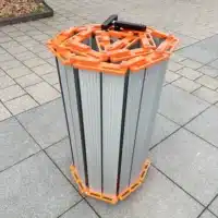 Eine zusammengerollte Rollrampe aus Aluminium mit orangefarbenen Seitenteilen steht auf einer gefliesten Außenfläche.